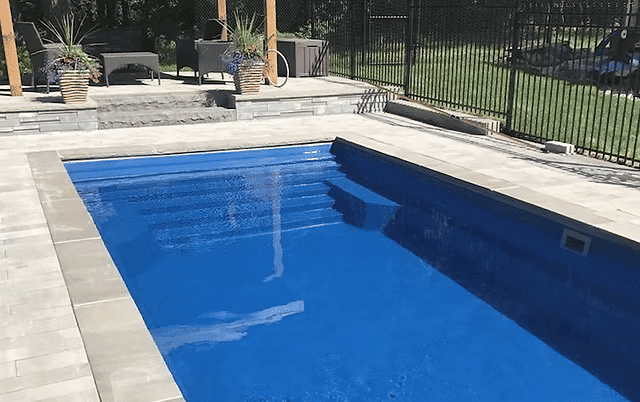 Inground swimming pool with steps, surrounded by landscaping and a fenced yard.